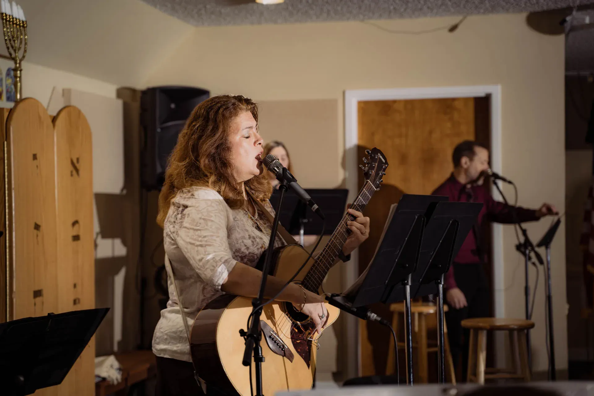 A woman with auburn hair plays acoustic guitar and sings into a microphone with eyes closed; behind her, two more singers lead worship at music stands beside the carved wooden ark doors.
